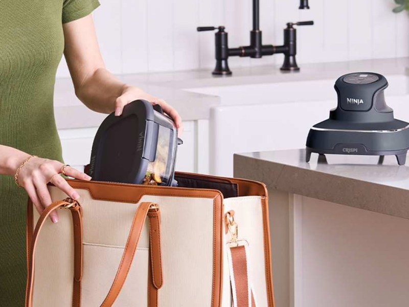 Eine Person packt einen Behälter mit Essen in eine Handtasche. Im Küchenhintergrund steht der Deckel der neuen Ninja Crispi Heißluftfritteuse.